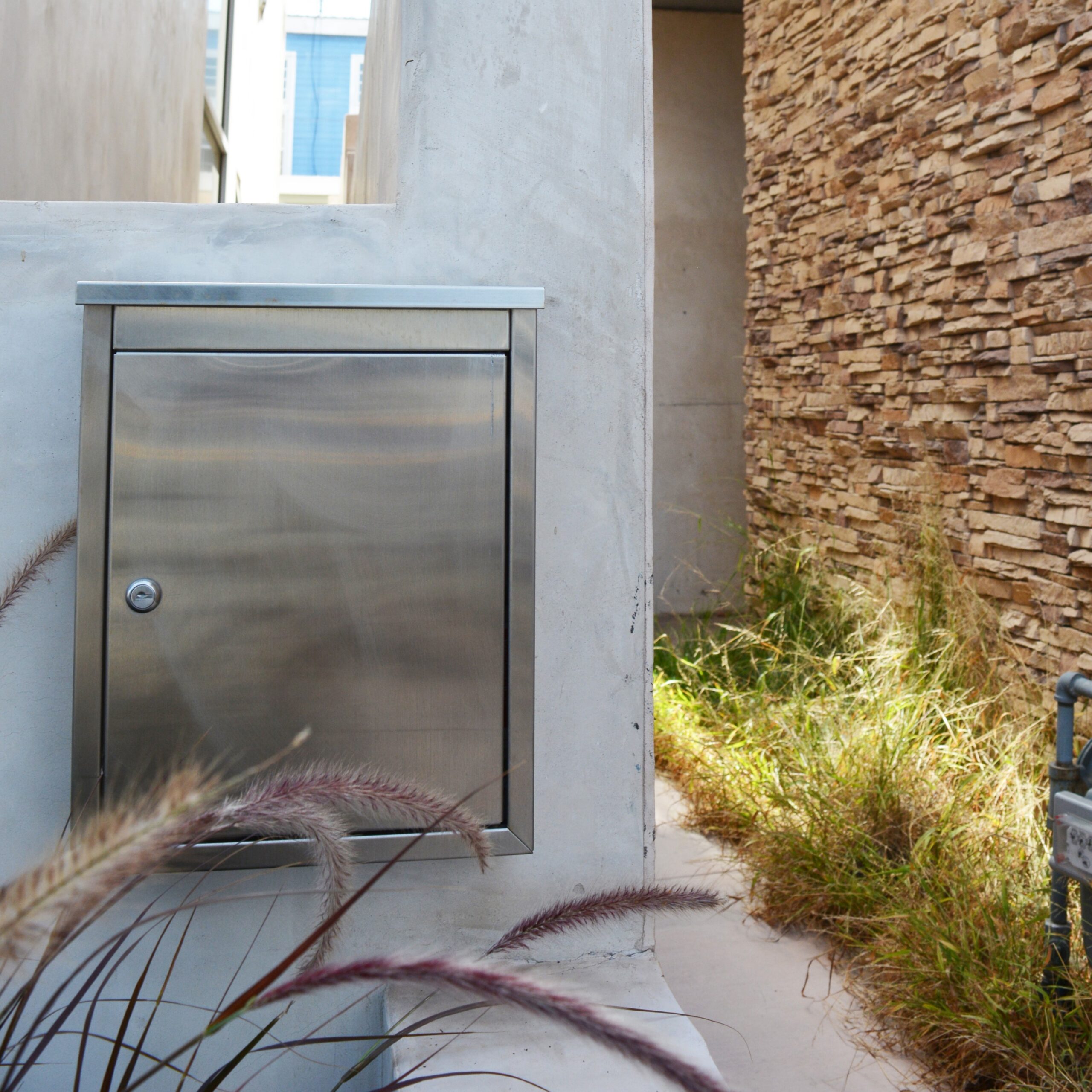 Stainless metal mailbox or cabinet mounted on a concrete wall beside a stone accent wall, with ornamental grasses in the foreground.