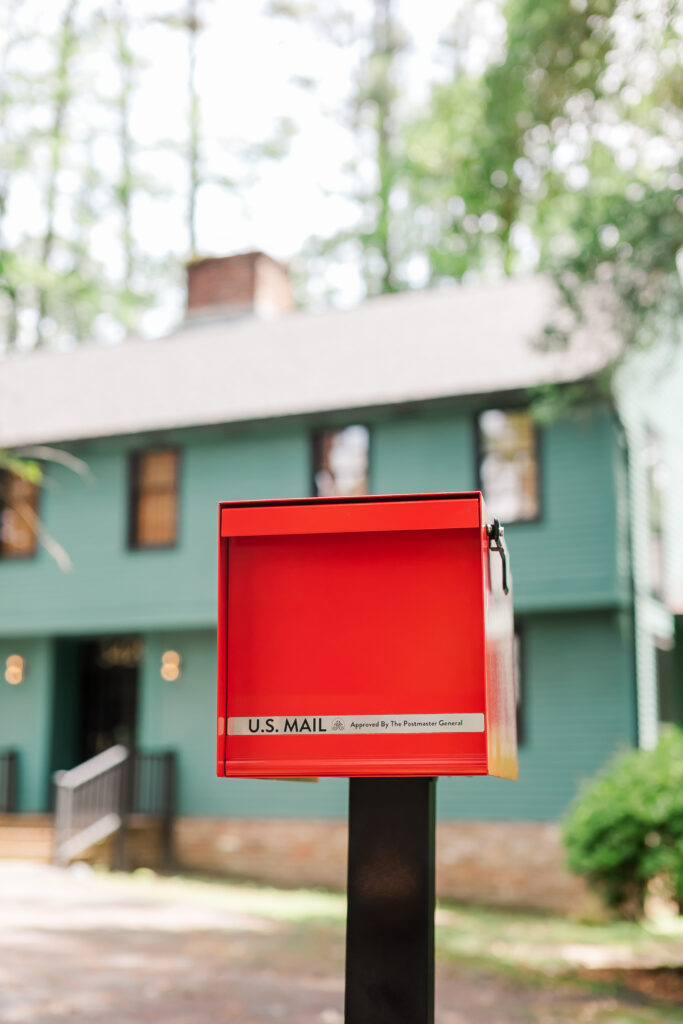 Red U.S. Mail mailbox on a black post in front of a green house, with trees blurred in the background, indicating a street scene for sending letters.