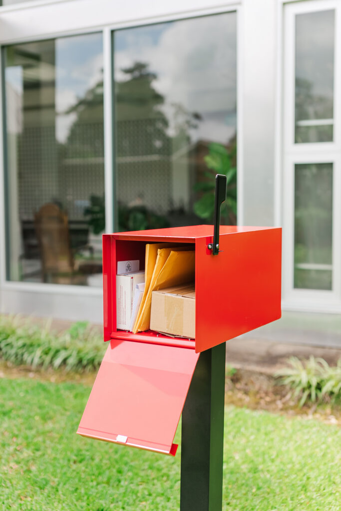 Red mailbox on a dark post outside, open with mail and packages visible inside on a green lawn in front of a modern house