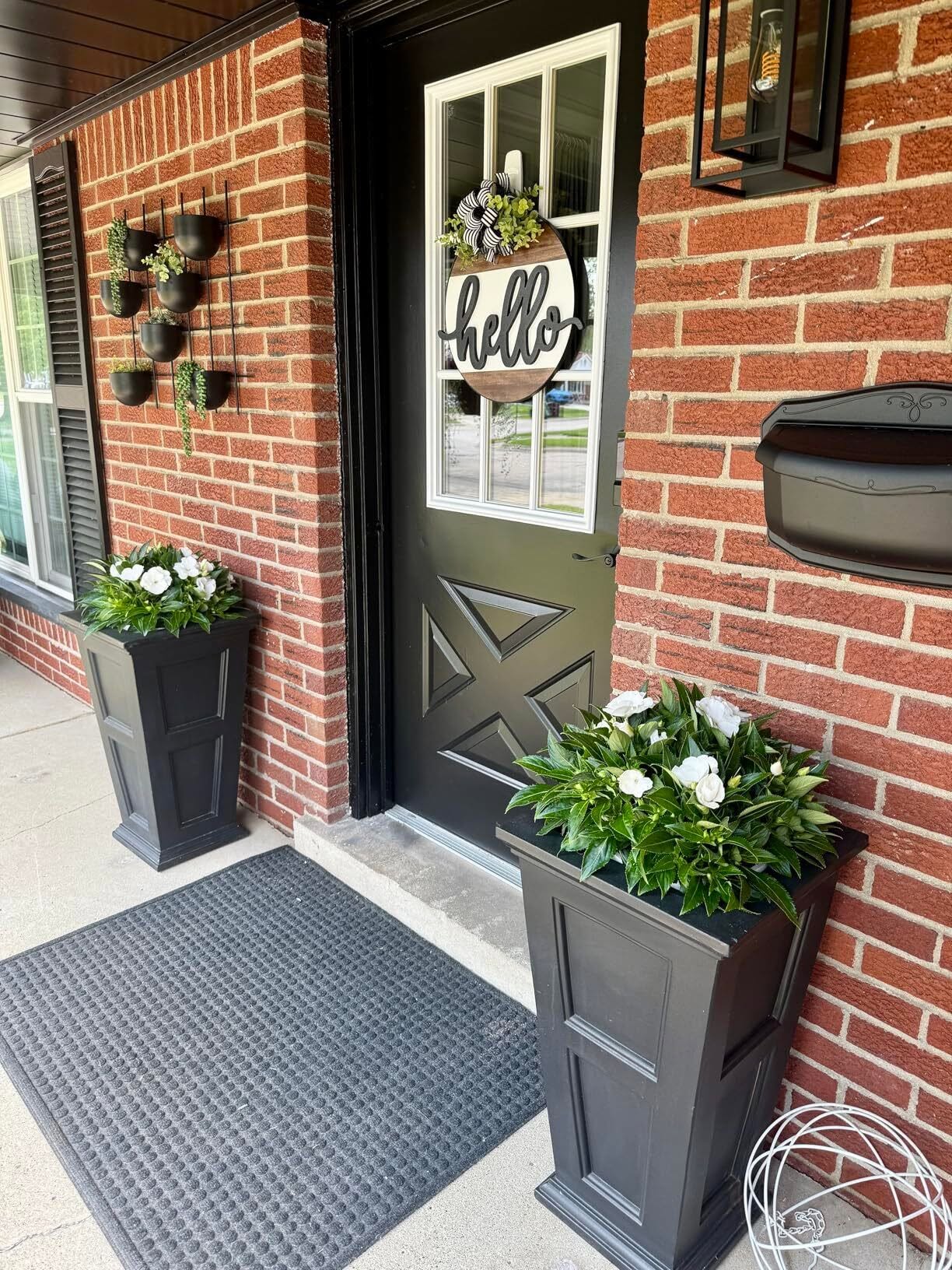 Black front door with a round wooden 'hello' sign and greenery, flanked by black planters with white flowers on a brick porch.