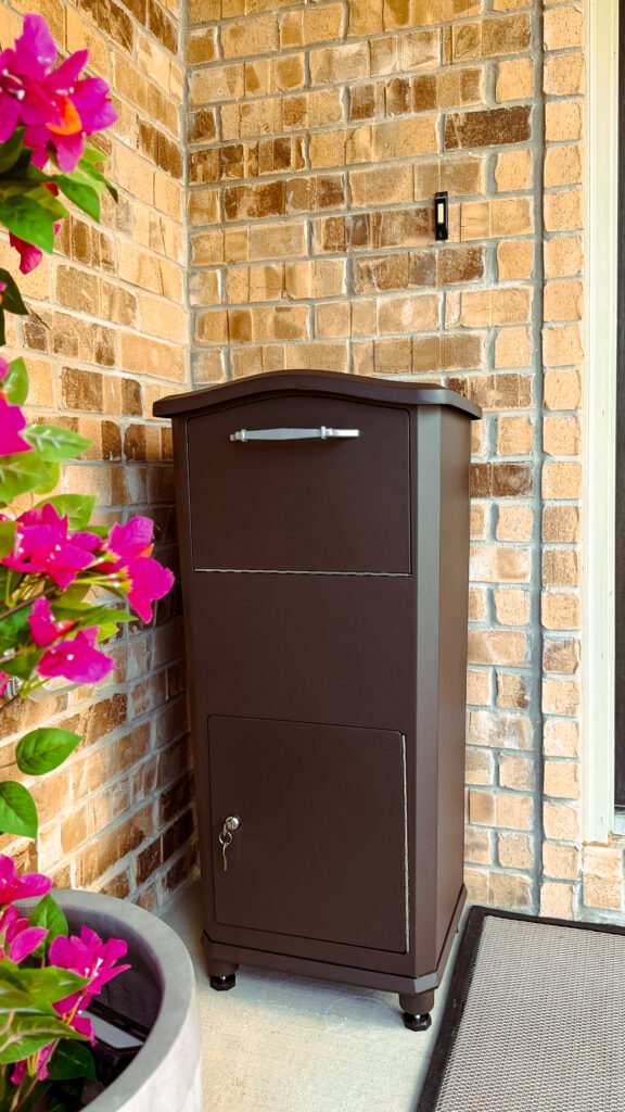 Brown metal outdoor mailbox with a curved top, set against a brick wall; has a small key lock on the lower door, and vibrant pink flowers appear on the left.