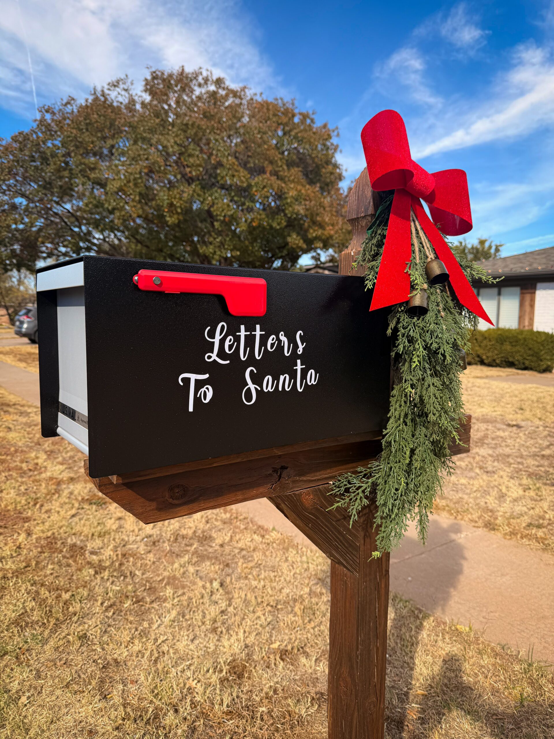 Black mailbox with a red flag and a festive red bow, labeled 'Letters to Santa' on a wooden post in a yard.