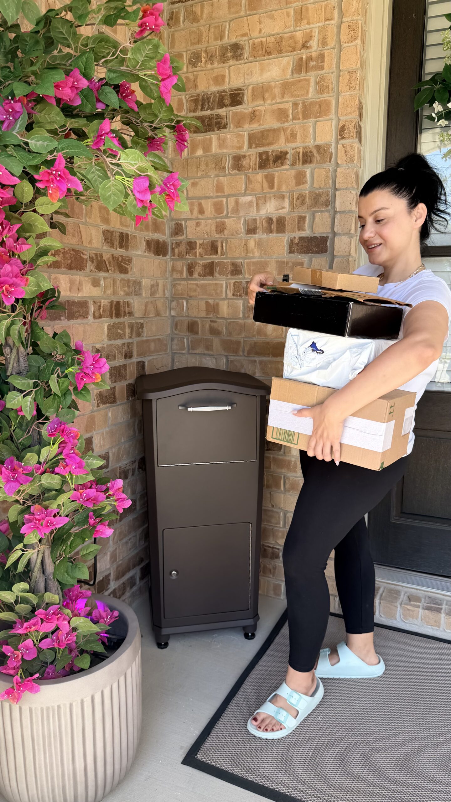 Delivery person on a home porch carrying several packages beside a tall dark mailbox, with pink bougainvillea flowers and a brick wall in the background.
