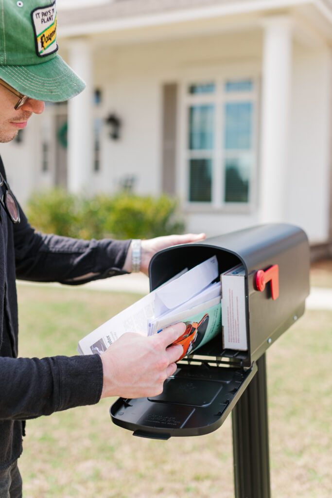 A person wearing a green cap deposits papers into a residential mailbox outdoors.
