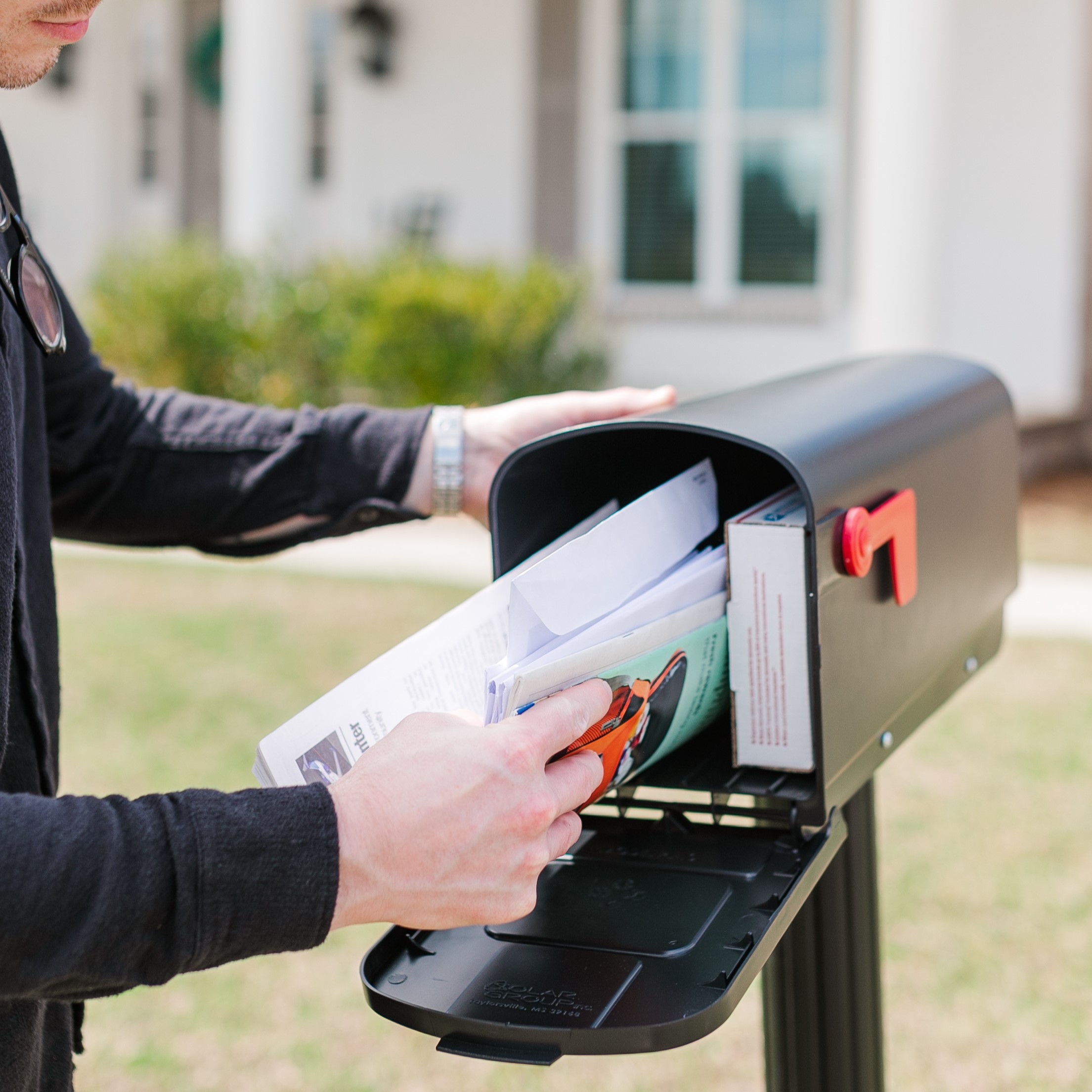 Person inserts mail into a black outdoor mailbox, hands visible holding letters and envelopes of various colors.