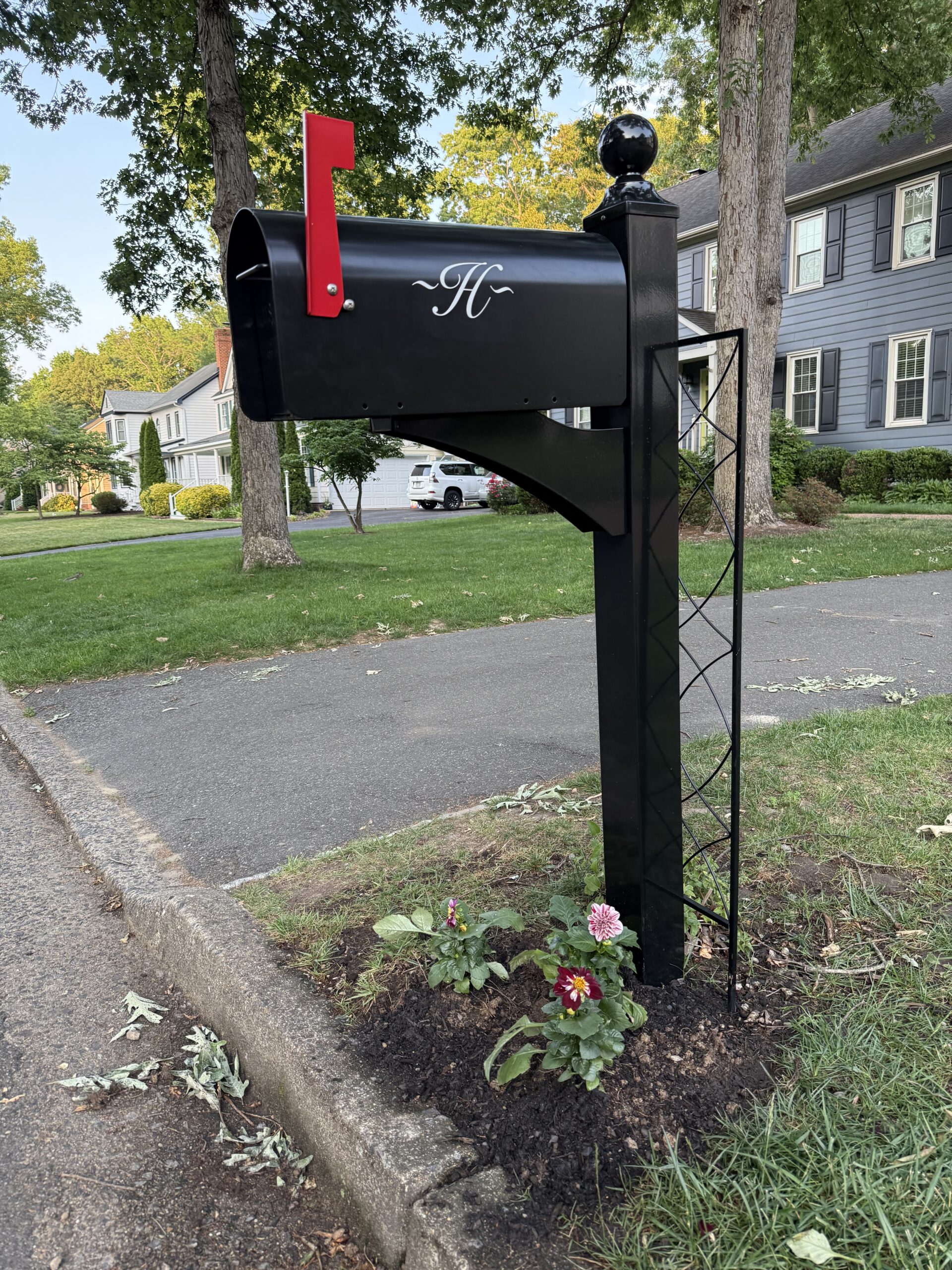 Black mailbox with red flag mounted on a tall post in a suburban yard; flowers planted at the base.