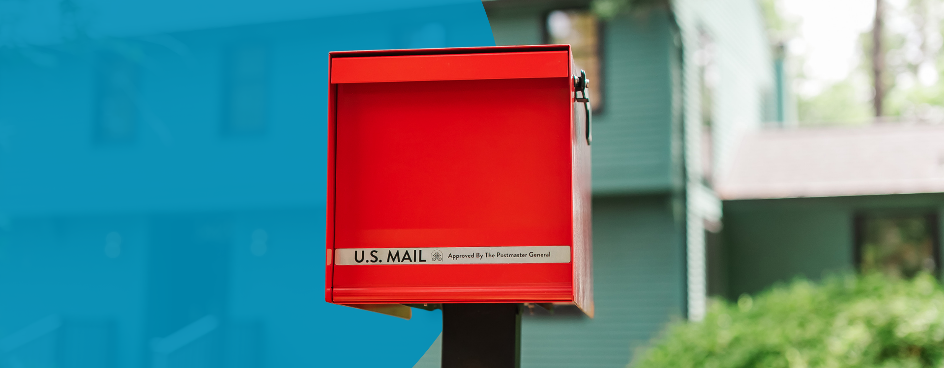 Red U.S. mailbox on a post beside a green house in a suburban setting.