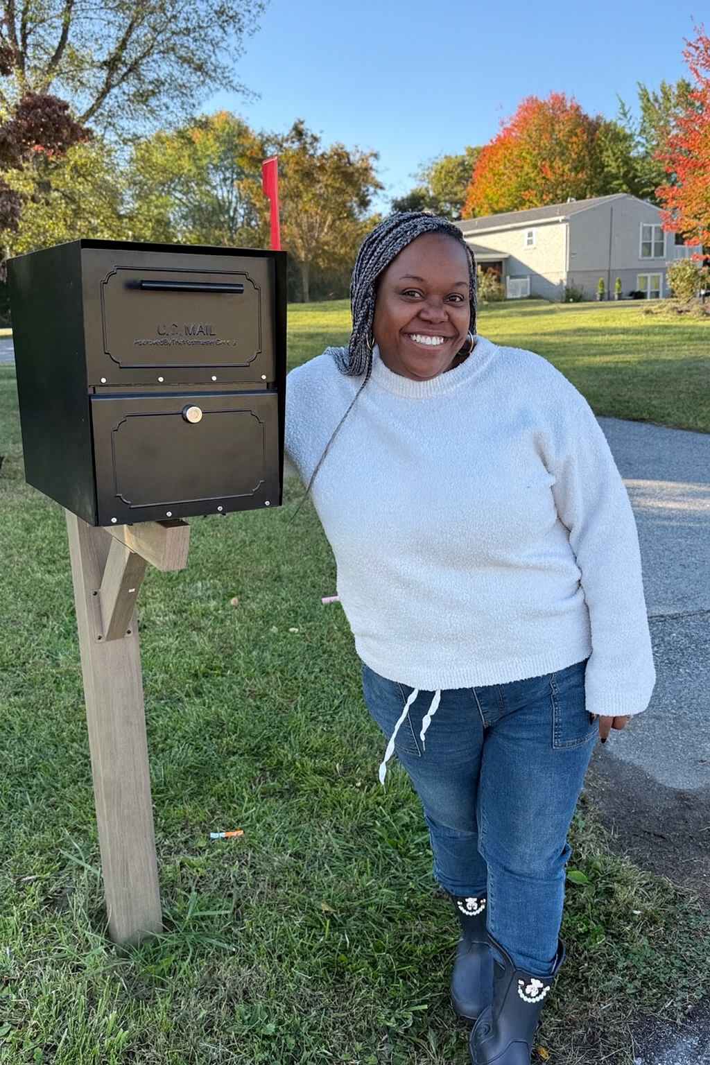 Smiling woman in a white sweater stands beside a black mailbox on a wooden post in a suburban yard with fall trees in the background.