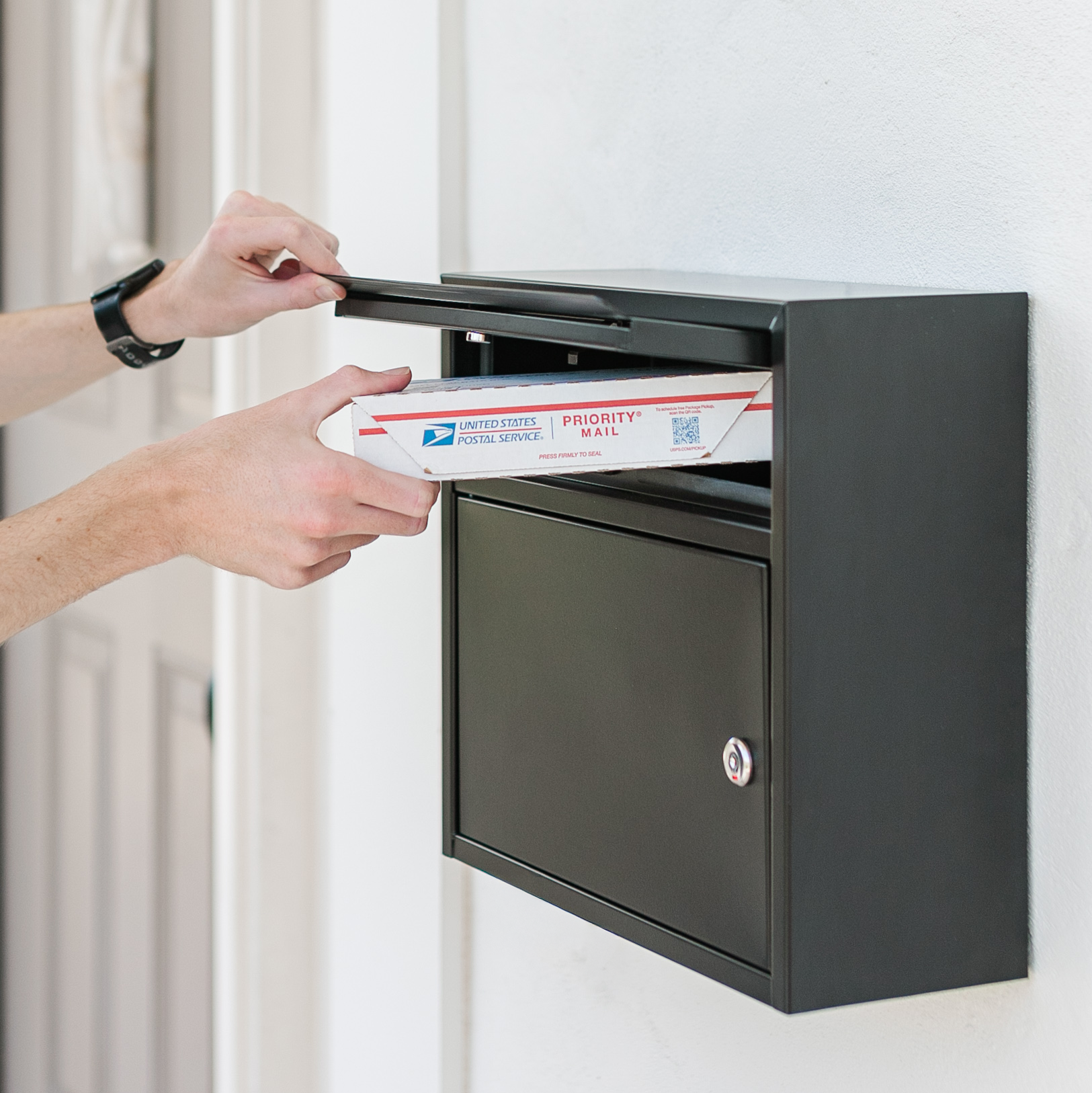 Person placing a USPS Priority Mail envelope into a black wall mailbox at a doorway with a lantern above and a green wreath in the background