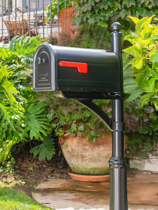 Black US mailbox on a post with the red flag raised, set among lush garden plants and a large planter.