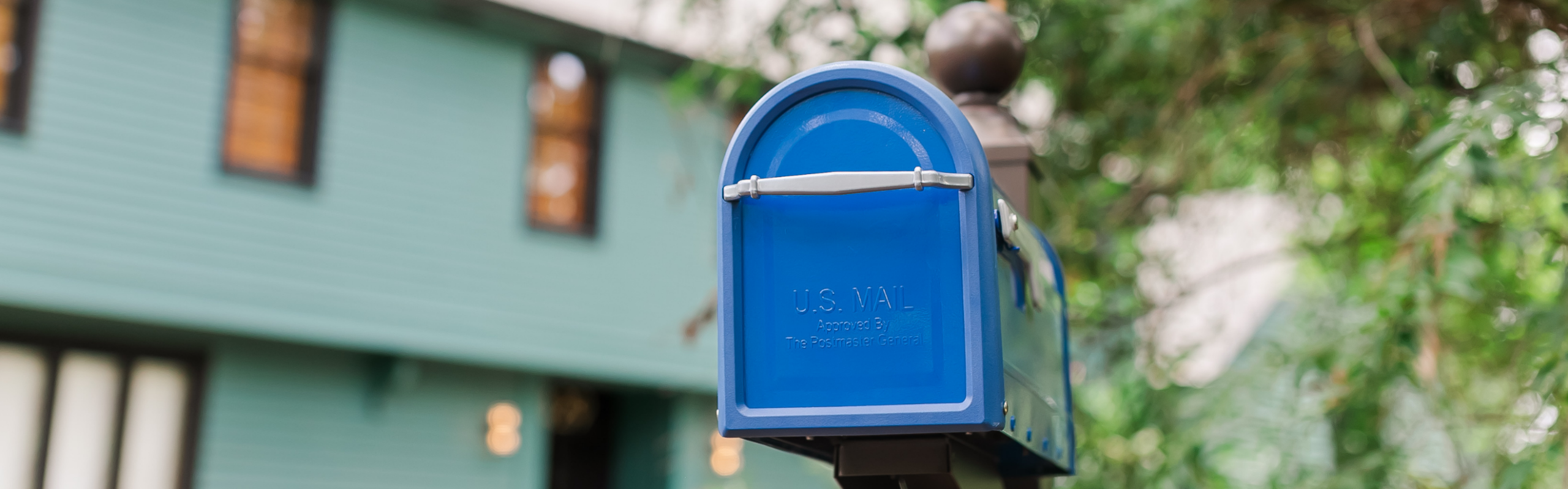Blue U.S. Mail mailbox on a post along a residential street, with a green house blurred in the background.