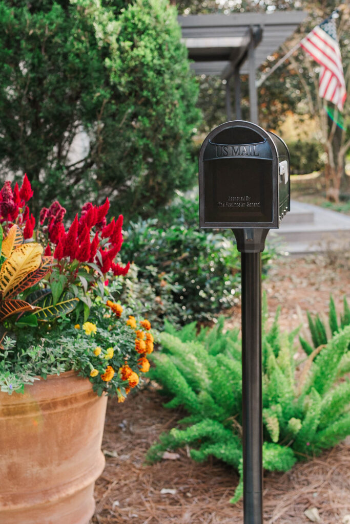 Black US Mail mailbox on a post beside a flower bed with red and orange blooms and greenery, American flag in background