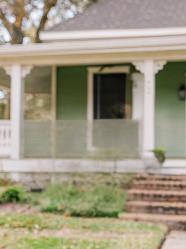 Blurry image of a green front porch with white columns, railing, and brick steps leading up to the entryway.