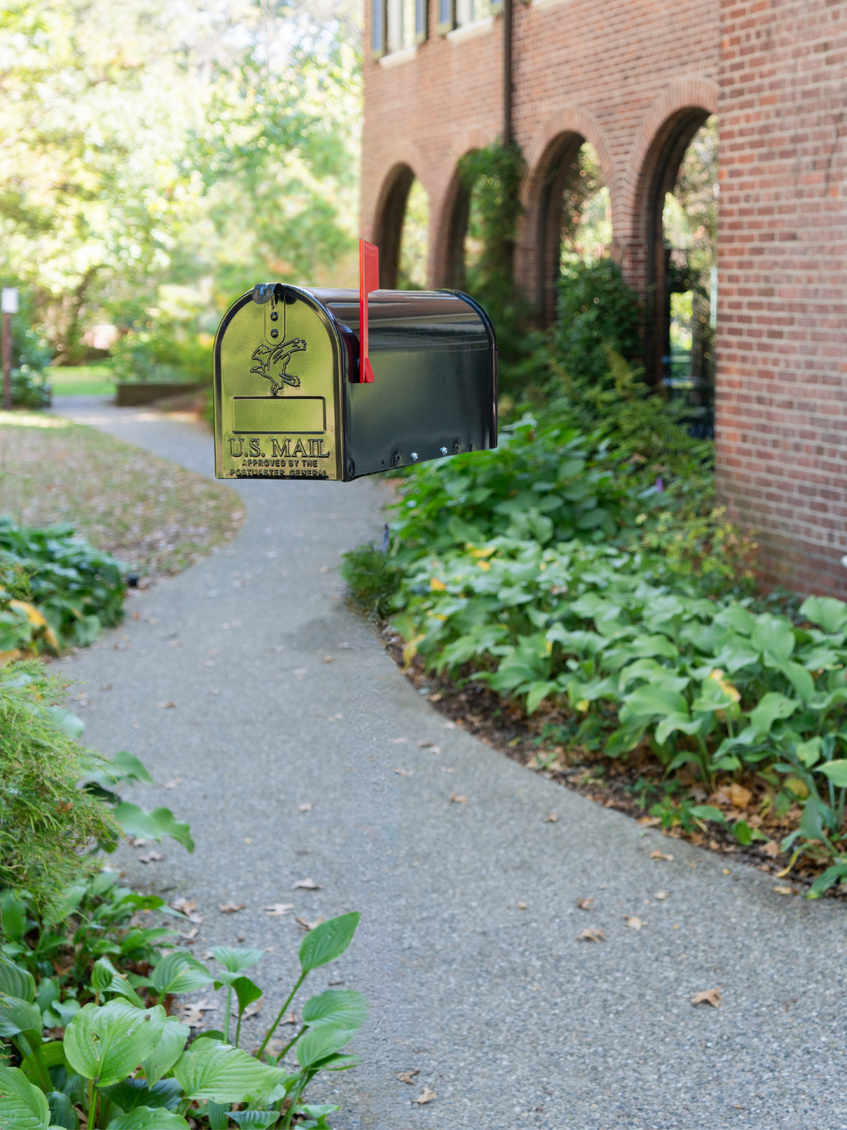 Suspended black metal US Mailbox with a red flag raised, along a brick building and leafy walkway.