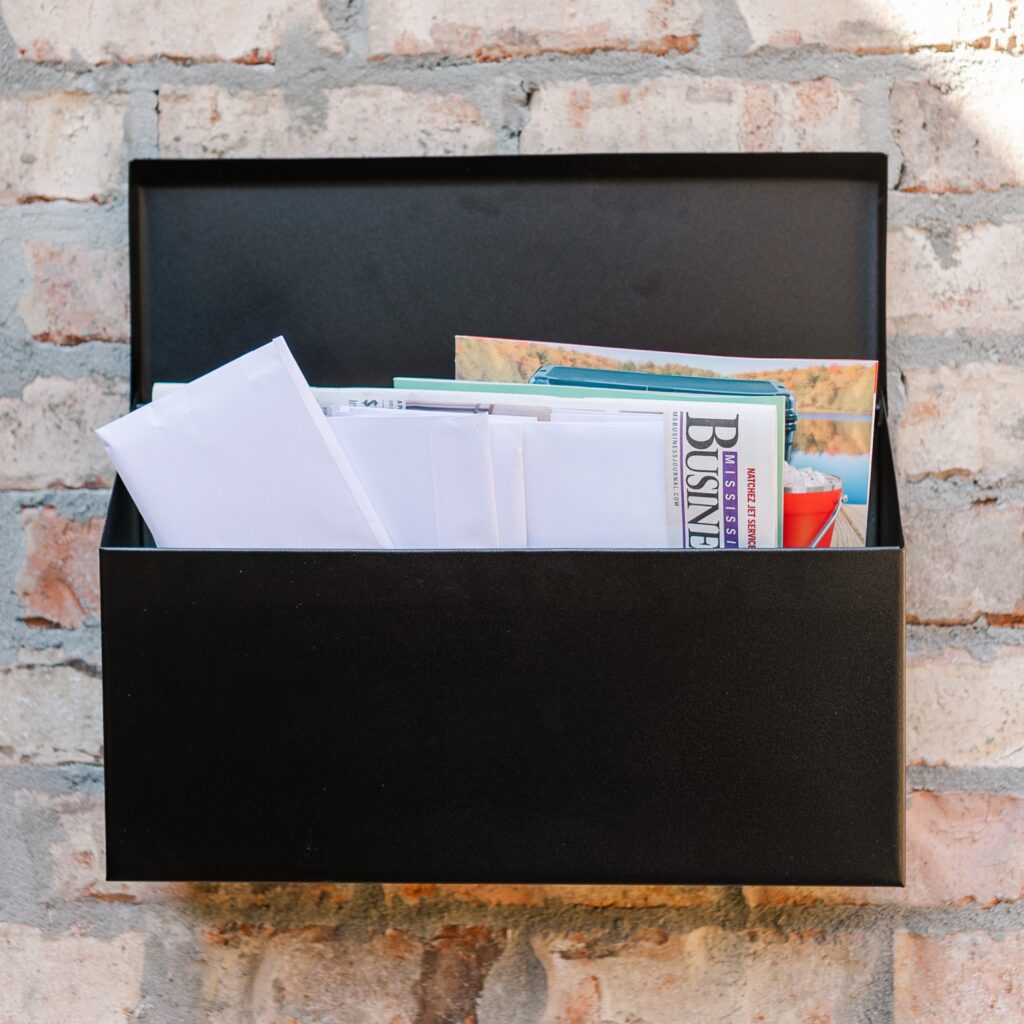 Open black metal mailbox with letters and magazines protruding against a textured brick wall, conveying mail delivery.
