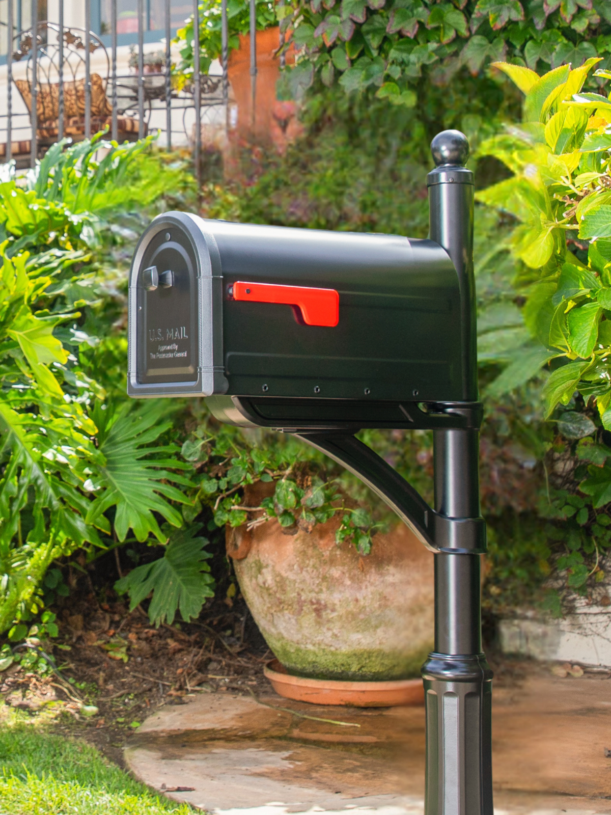 Black U.S. Mail mailbox with a red flag on a dark post beside lush greenery in a residential yard.