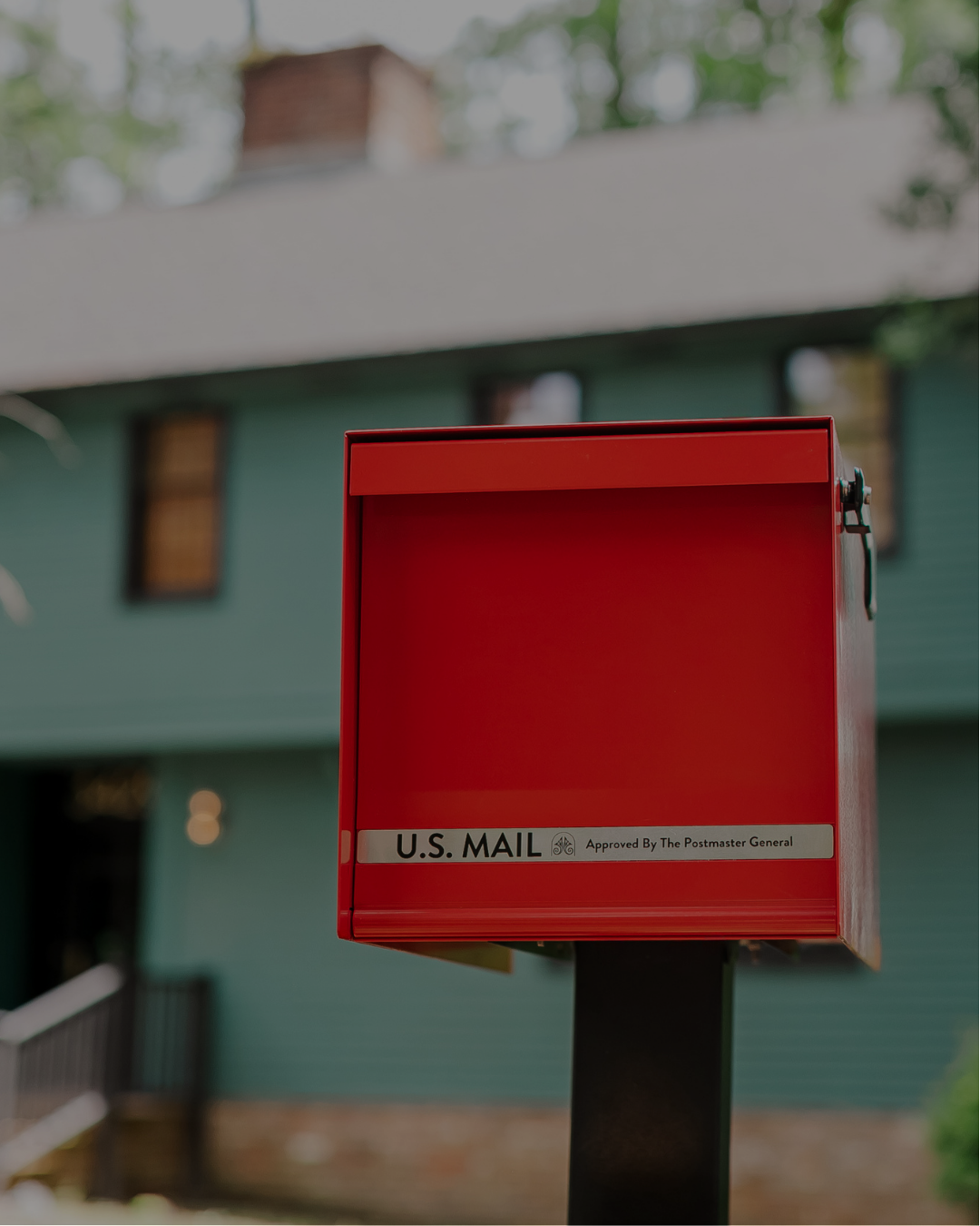 Red U.S. Mail mailbox on a black post in front of a blurred green house.