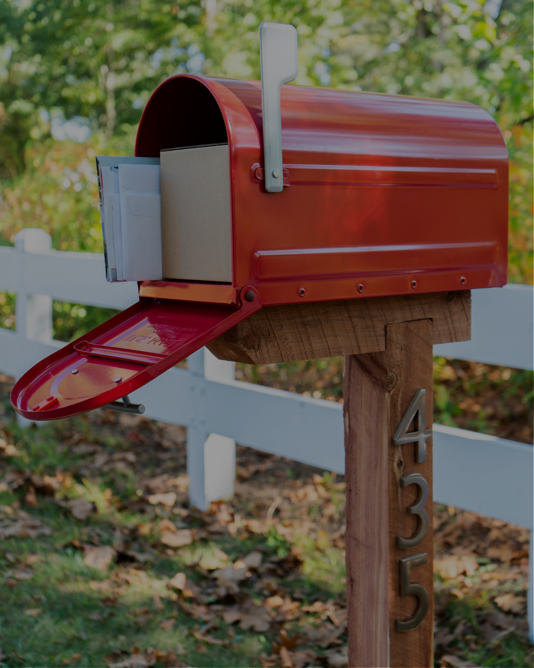 Red mailbox on a wooden post with its door open and mail inside, beside a white fence and fallen leaves.
