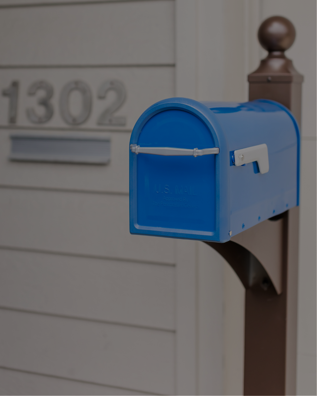 Blue U.S. Mail mailbox mounted on a post outside a house, with address 1302 visible in the background.