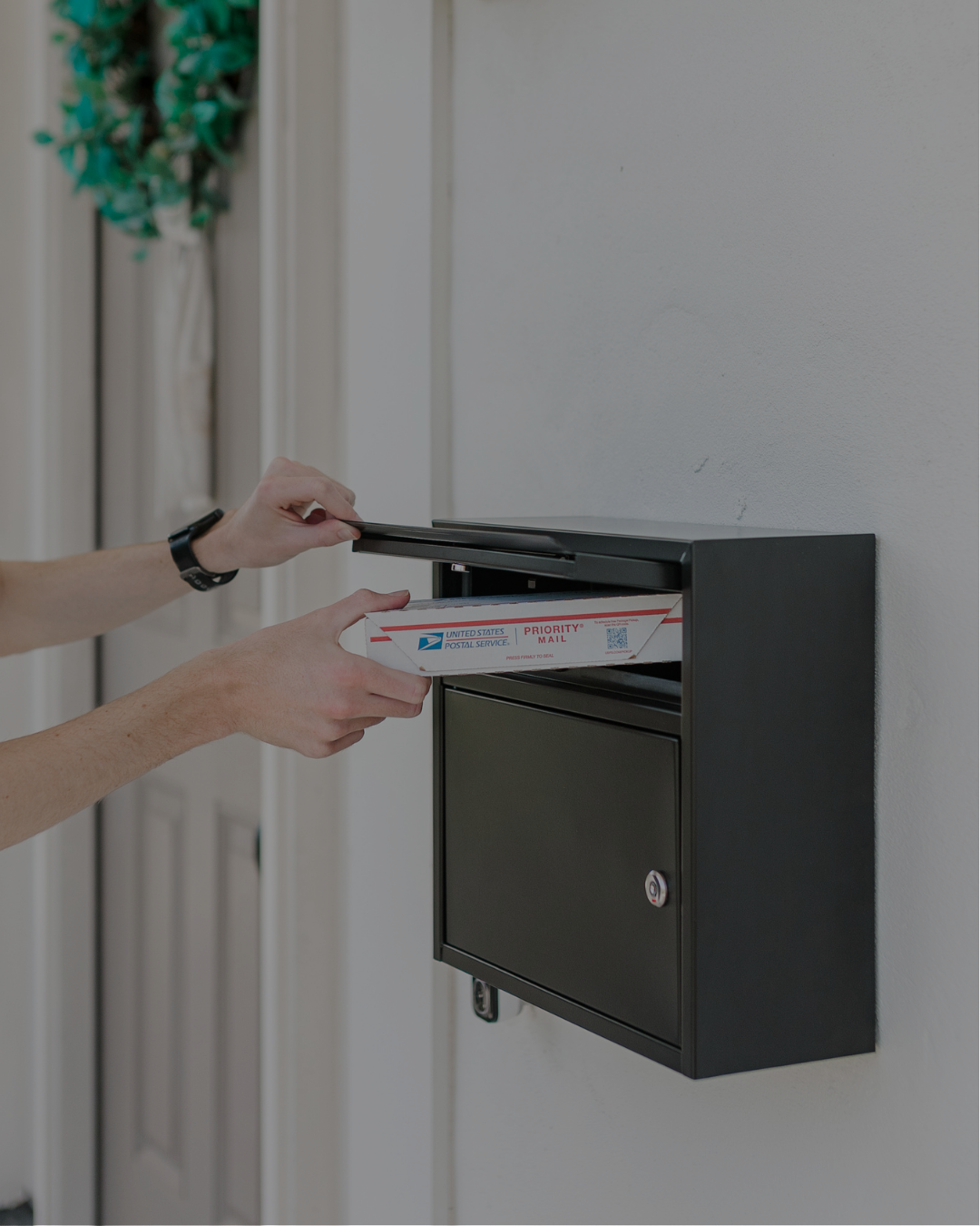 Person inserts a USPS Priority Mail box into a wall-mounted mailbox at an apartment entrance.