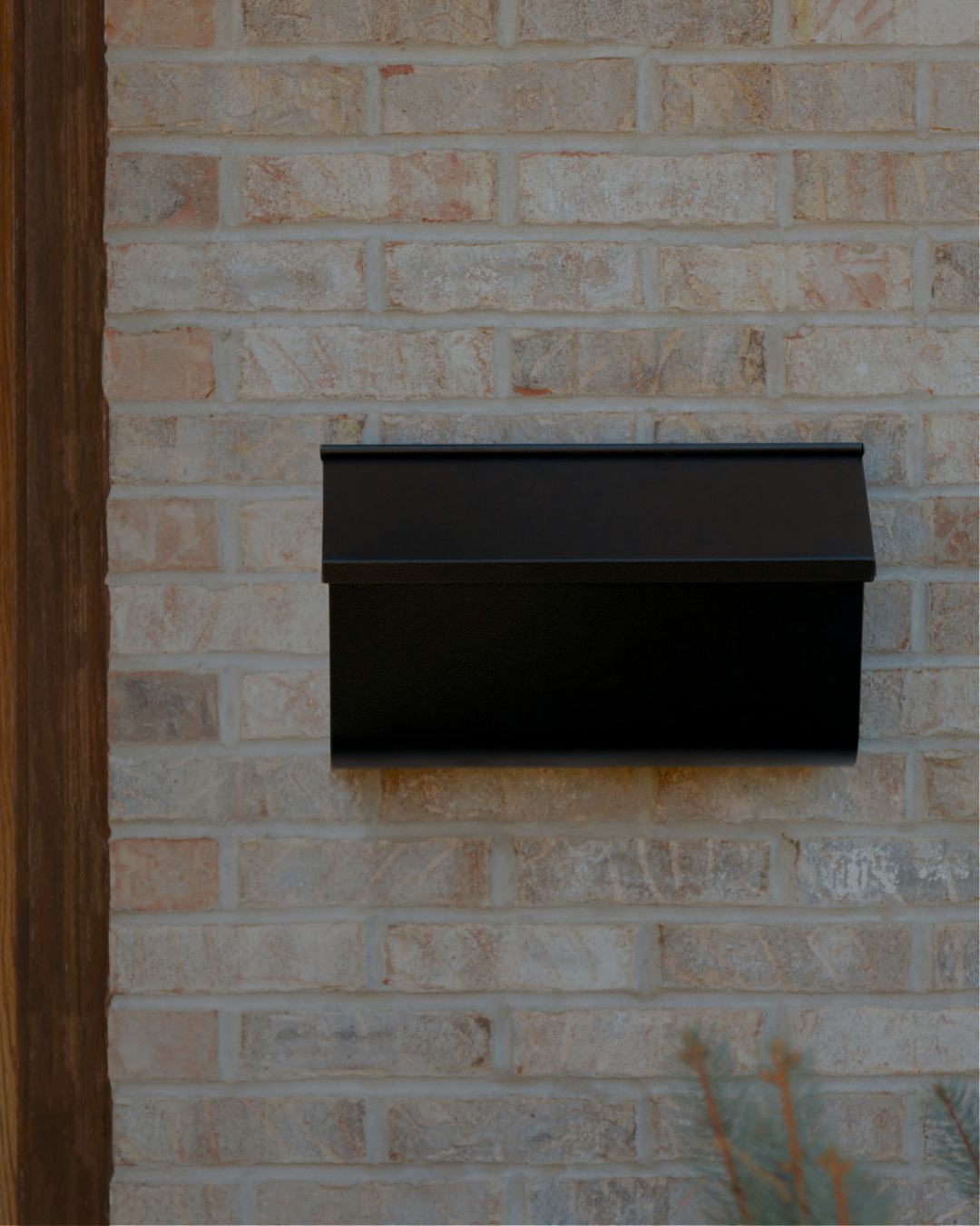 Black metal mailbox mounted on a light brick exterior wall, with a wooden post on the left.”],